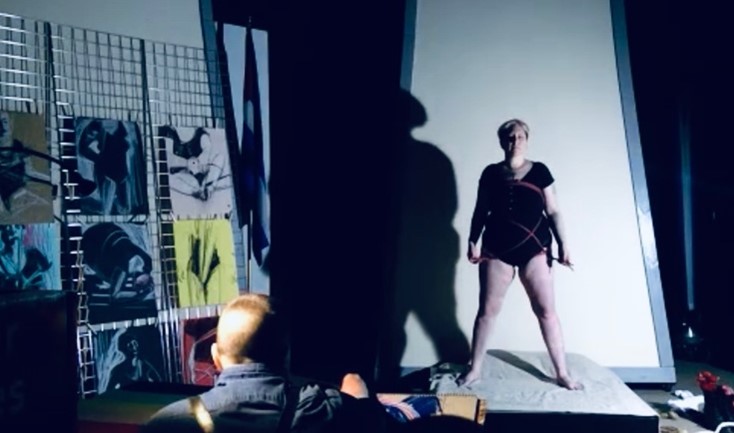 The image shows an art studio with a few drawings on the left side, a painter in the middle and a non-binary person stand in front of the painter. The performer is a white person with a blond short hair and is using a black outfit with short sleeves and black short pants. 
