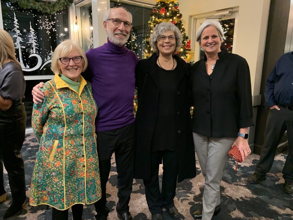 Four college deans standing in front of a Christmas tree. From left to right: Dr. Virginia Cooke, Dr. Eric Davis, Dr. Jacqueline Nolte, and Dr. Sylvie Murray. Dr. Cooke is dressed in a vibrant green and yellow patterned jacket, wearing glasses with short, straight hair. Dr. Davis is wearing a purple sweater and black pants, complemented by glasses. Dr. Nolte is seen in a stylish black jacket, blouse, and pants, with glasses and short, curly hair. Dr. Murray, wearing a black blouse, black pants, and a short, straight hair. The group stands closely together and smiling.