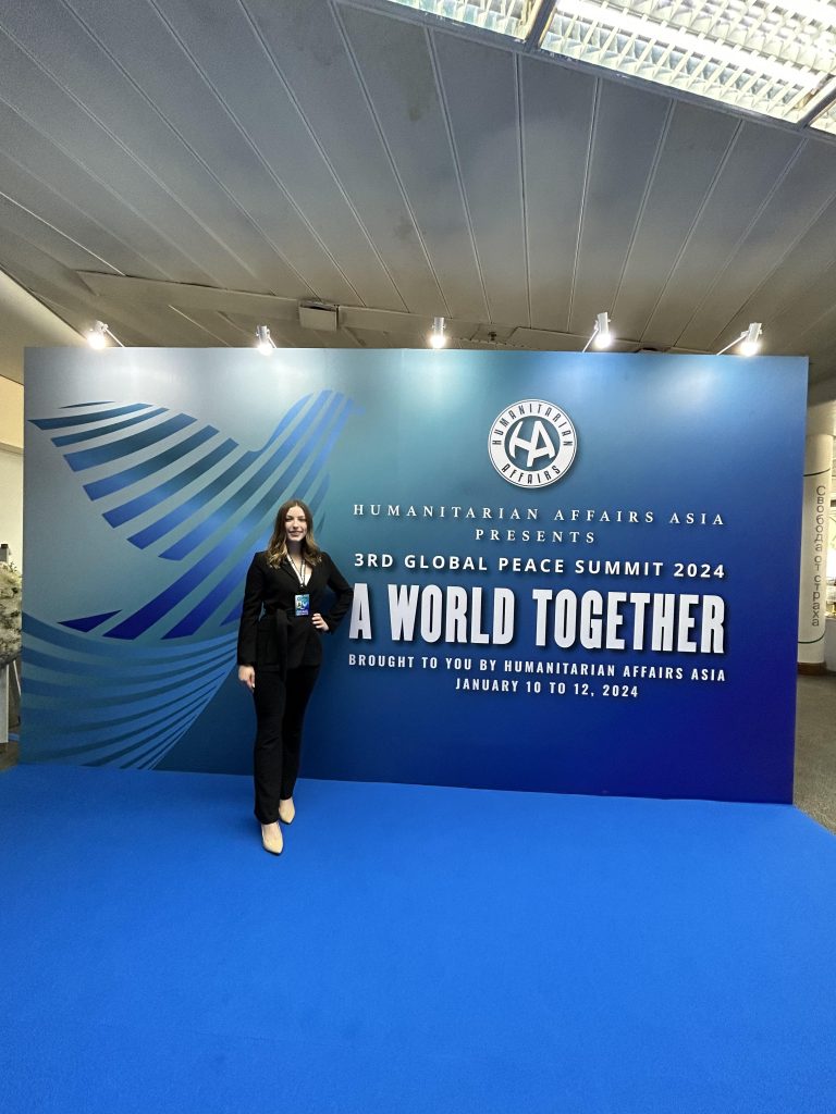 Ashley McDougall in front of a blue panel that reads "Humanitarian Affairs Asia presents: 3rd Global Peace Summit 2024 - A world together." Ashley is smiling, standing and wearing a black suit.