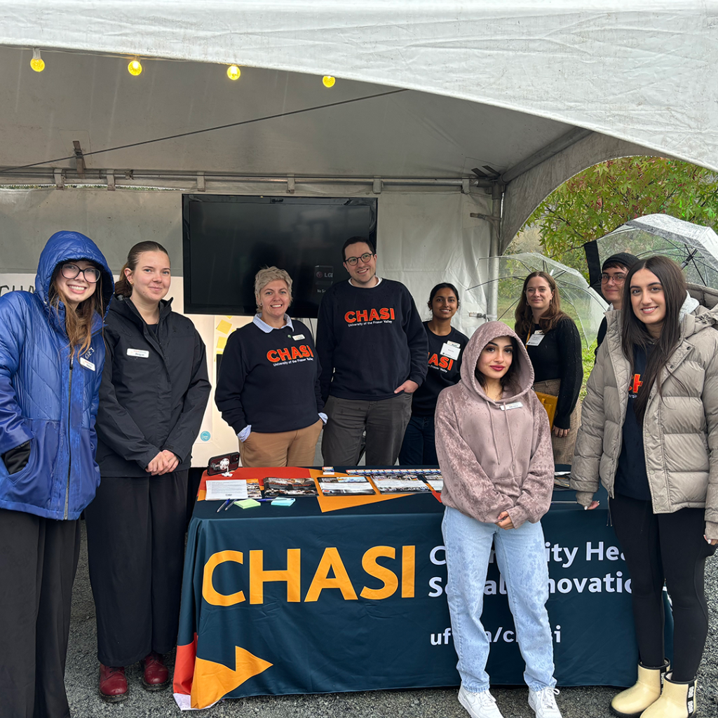 Photo of nine members of the CHASI team posing around CHASI's table at the event. They are all smiling for the camera, despite the rainy day.