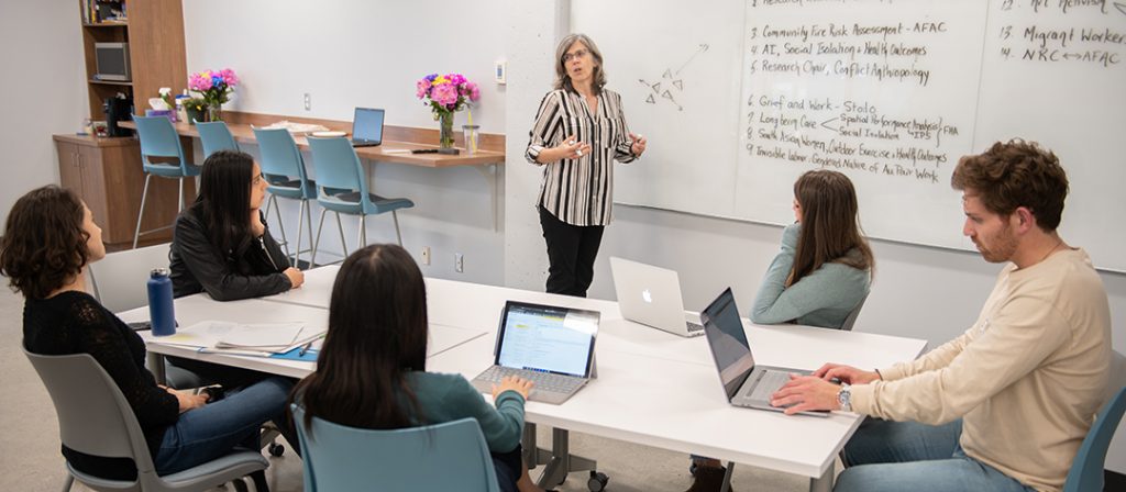 Martha Dow stands at a white board, speaking to the CHASI team who are seated around a table.