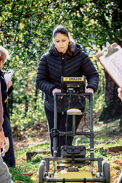 Photo of Dr. Sarah Beaulieu pushing a Ground Penetrating Radar machine in a wooded area, while people with clipboards observe and take notes.