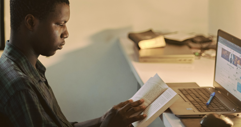 Photo of a young person sitting at their open laptop, studying a book with a pen resting on their keyboard.