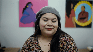 An animated GIF of Celina Koops sitting in front of her artwork, tilting and bowing her head with a smile