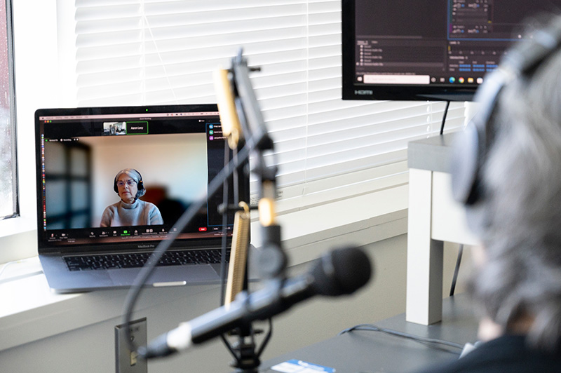 Photo of a studio with professional recording equipment. The focus is on a laptop showing Dr. Jacqueline Nolte on a Zoom call, while in the foreground, Dr. Martha Dow speaks into a microphone.