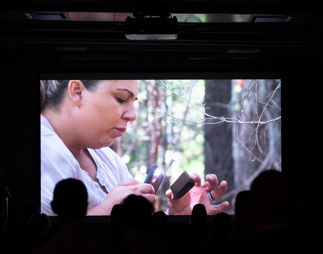 A video of Dr. Sarah Beaulieu examining a small artefact in a forest plays on a large movie theatre screen, with silhouetted heads in the foreground