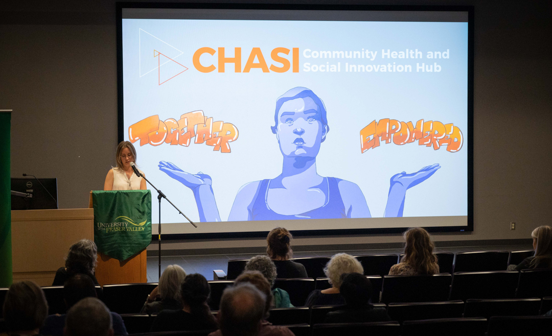 Ekat Marenkov addresses an audience in a theatre, while a screen behind her shows the CHASI hub's logo and the "together empowered" mural on the Hub's wall.