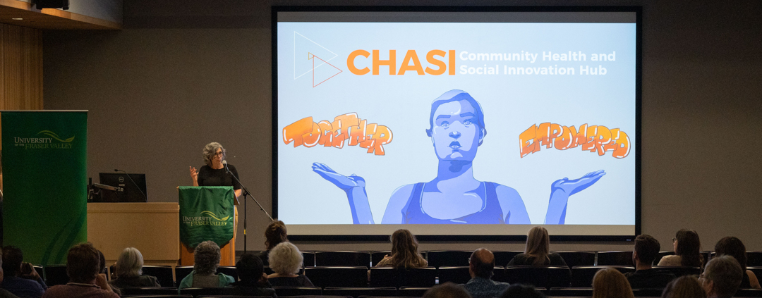 Dr. Martha Dow addresses an audience in a theatre, while a screen behind her shows the CHASI hub's logo and the "together empowered" mural on the Hub's wall.