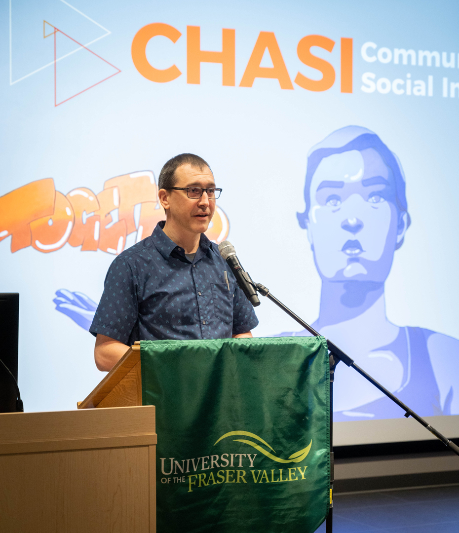 Greg Laychak addresses an audience in a theatre, while a screen behind her shows the CHASI hub's logo and the "together empowered" mural on the Hub's wall.