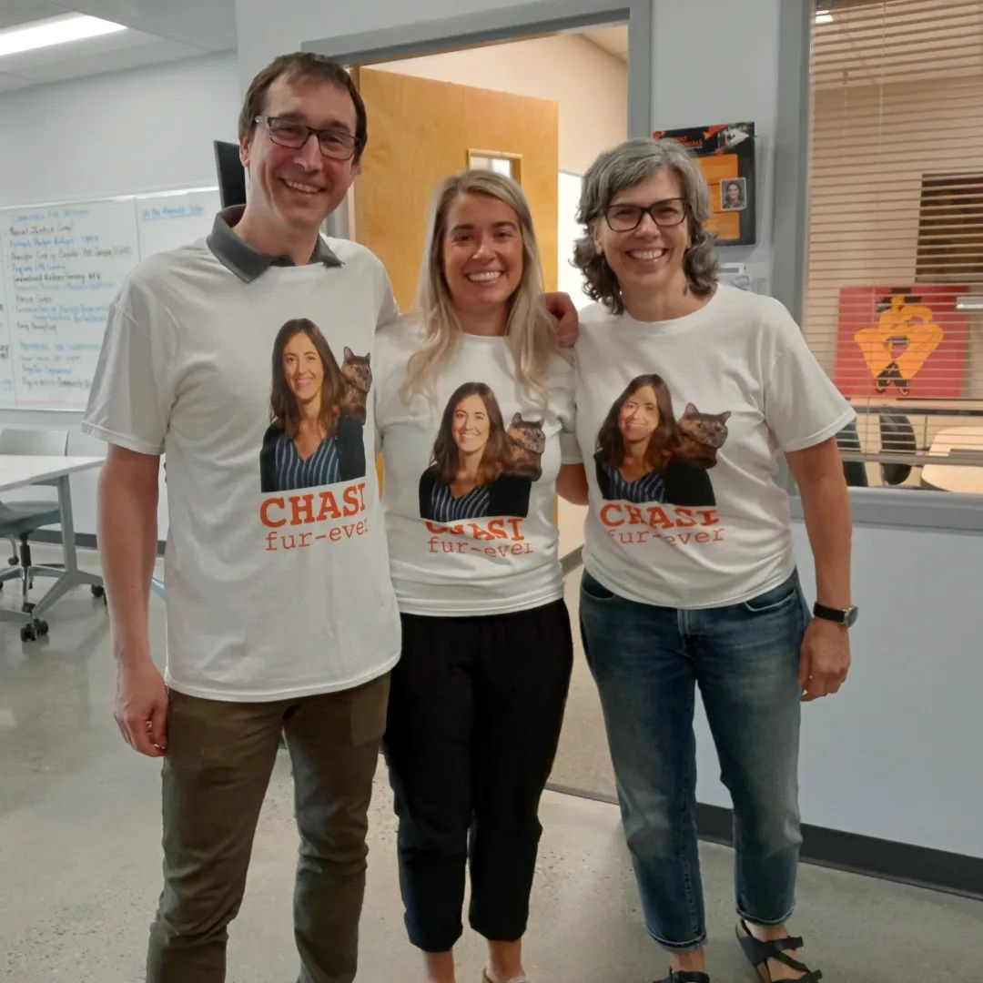 Photo of CHASI’s Greg Laychak, Larissa Kowalski, and Martha Dow, posing for a photo in the Hub while wearing custom t-shirts that read “CHASI fur-ever", with a photo of Esther on them and a cat on her shoulder. 