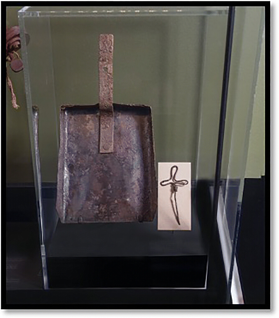 Photo of a museum display. Inside a transparent box is a rusted metal shovel head, and a small cross, formed out of a piece of barbed wire.