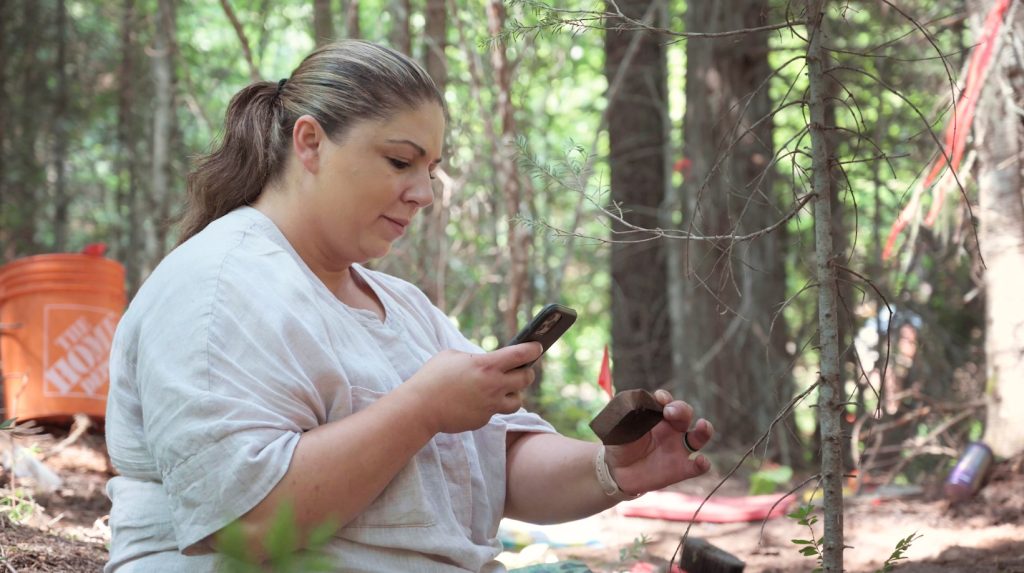 Photo of Dr. Sarah Beaulieu at a forested archaeological dig site, taking a photo of a small rusted tin with her phone.