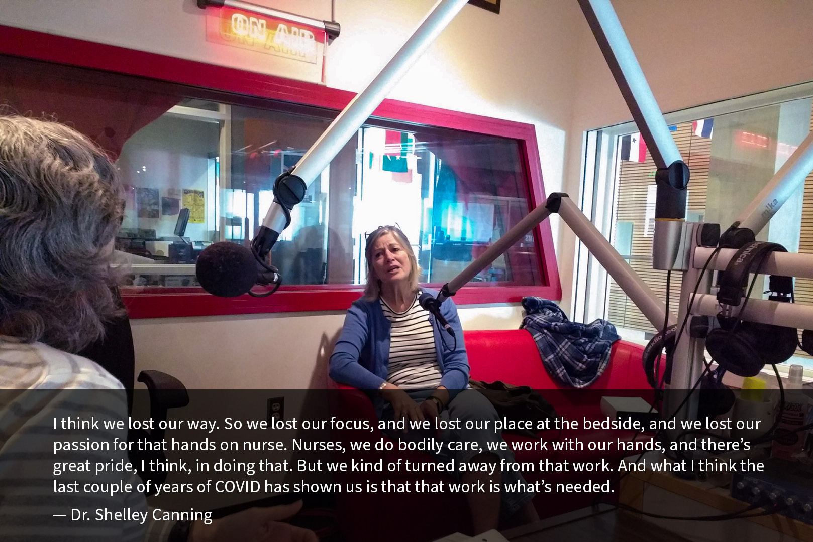 Dr. Shelley Canning sits on a couch in a radio studio opposite Dr. Martha Dow. They speak into microphones, with a glowing orange “on air” sign on the wall above Dr. Canning. A quote superimposed on the lower third of the image reads: “I think we lost our way. So we lost our focus, and we lost our place at the bedside, and we lost our passion for that hands on nurse. Nurses, we do bodily care, we work with our hands, and there’s great pride, I think, in doing that. But we kind of turned away from that work. And what I think the last couple of years of COVID has shown us is that that work is what’s needed.” The quote is attributed to Dr. Shelley Canning.