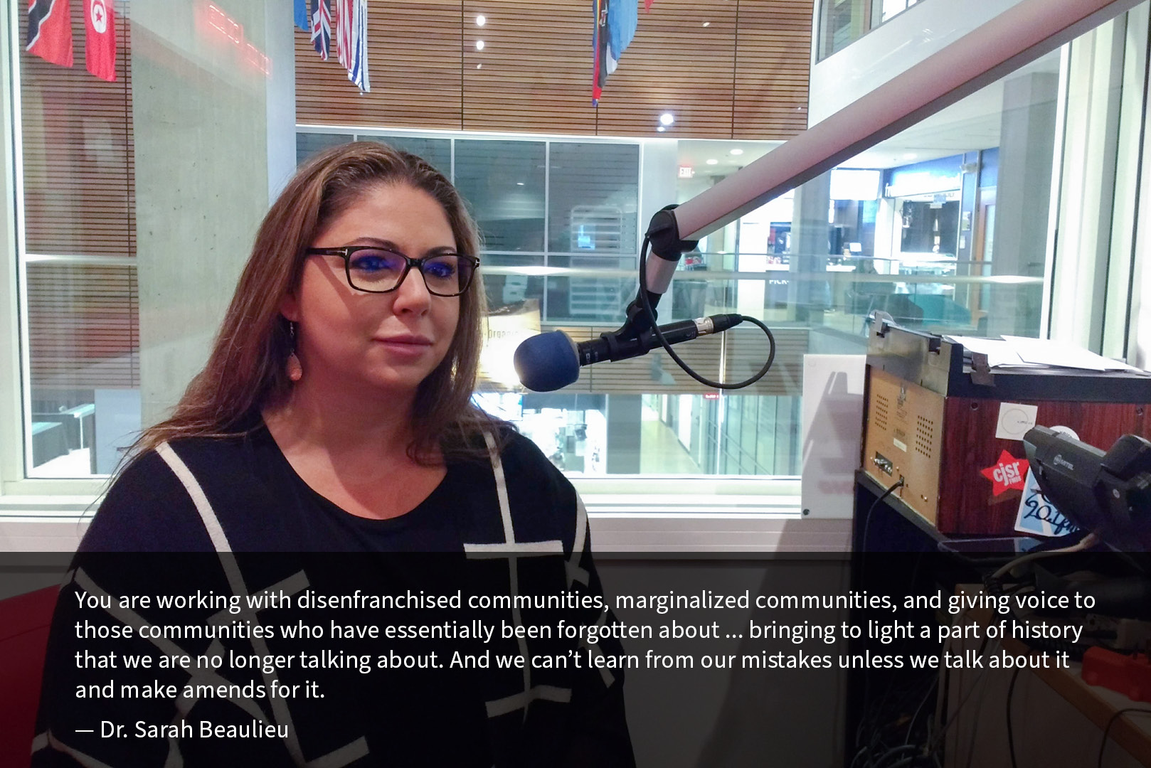 Dr. Sarah Beaulieu sits in the CIVL Radio studio with a microphone in front of her. Behind her, a window looks out at the UFV Abbotsford Student Union Building Atrium, with flags visible hanging from the ceiling above. A quote from Dr. Beaulieu reads: “You are working with disenfranchised communities, marginalized communities, and giving voice to those communities who have essentially been forgotten about ... bringing to light a part of history that we are no longer talking about. And we can’t learn from our mistakes unless we talk about it and make amends for it.”