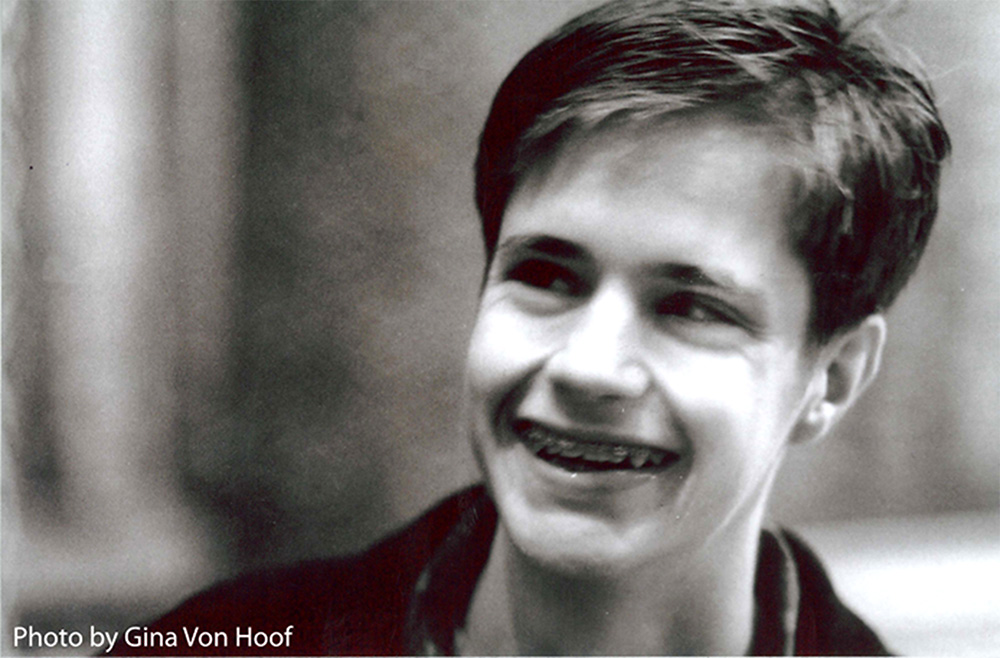 Black and white photo of Matthew Shepard, a young man in his early twenties with short dark hair. He is smiling or laughing, with braces on his teeth, and looking at something out of the frame. A credit on the photo reads: "photo by Gina Von Hoof"