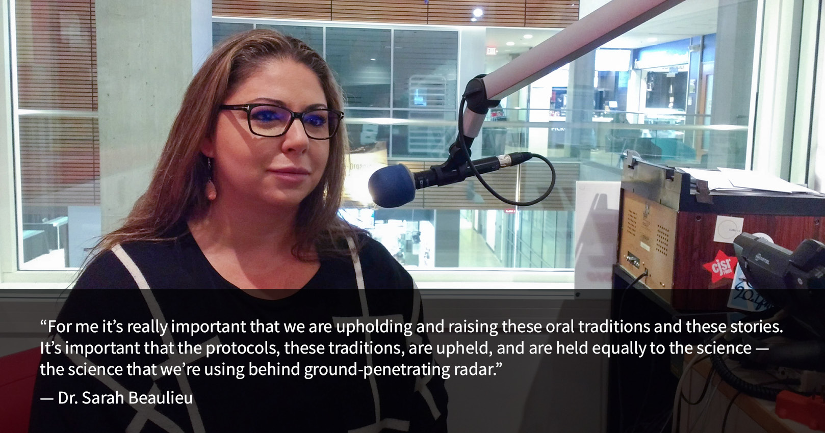 Dr. Sarah Beaulieu sits in the CIVL Radio studio with a microphone in front of her. Behind her, a window looks out at the UFV Abbotsford Student Union Building Atrium, with flags visible hanging from the ceiling above. A quote from Dr. Beaulieu reads: “For me it's really important that we are upholding and raising these oral traditions and these stories. It's important that the protocols, these traditions, are upheld, and are held equally to the science — the science that we're using behind GPR.”