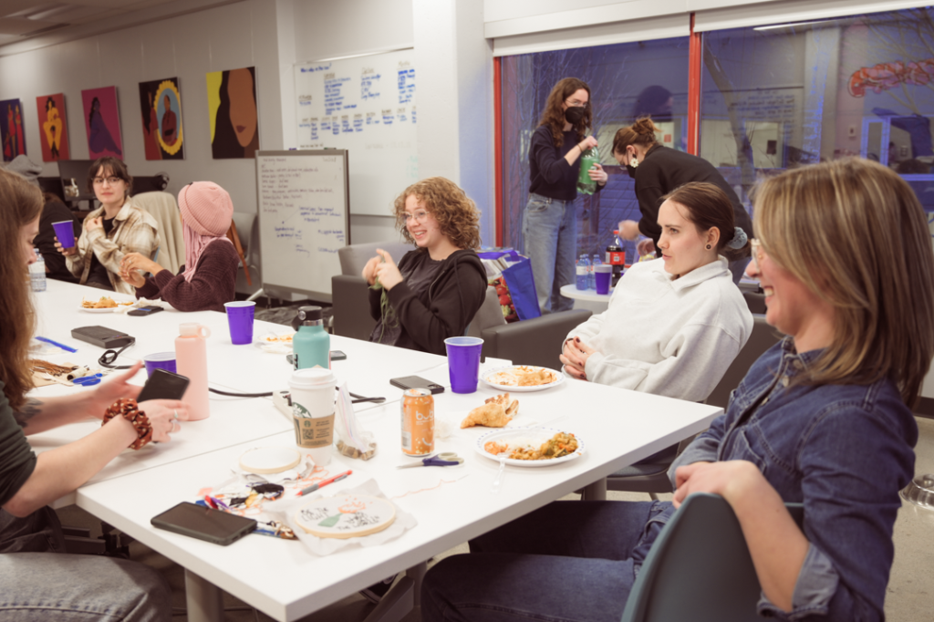 Photo of a group of women gathered around a large table eating, drinking, and talking. There are art supplies on the table.