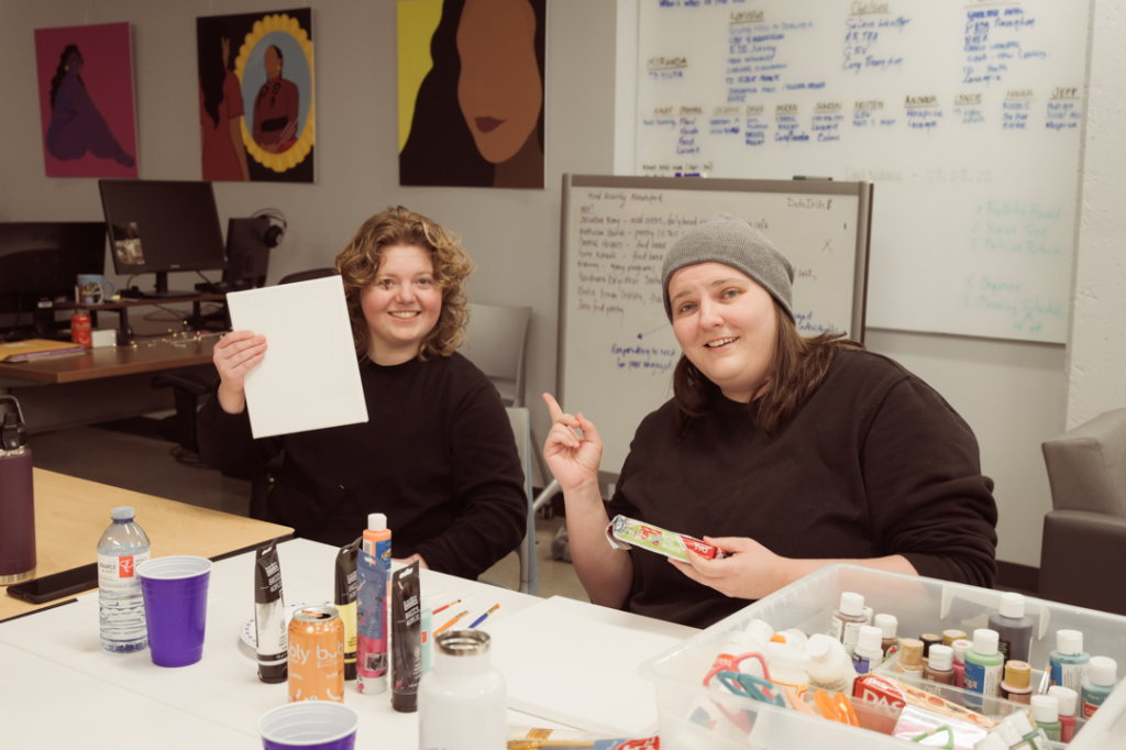 Photo of Kristen Bencze and Carlanna Thompson at a table filled with art supplies. Kristen is holding up what appears to be a blank piece of paper to show the camera, and Carlanna is pointing to it.