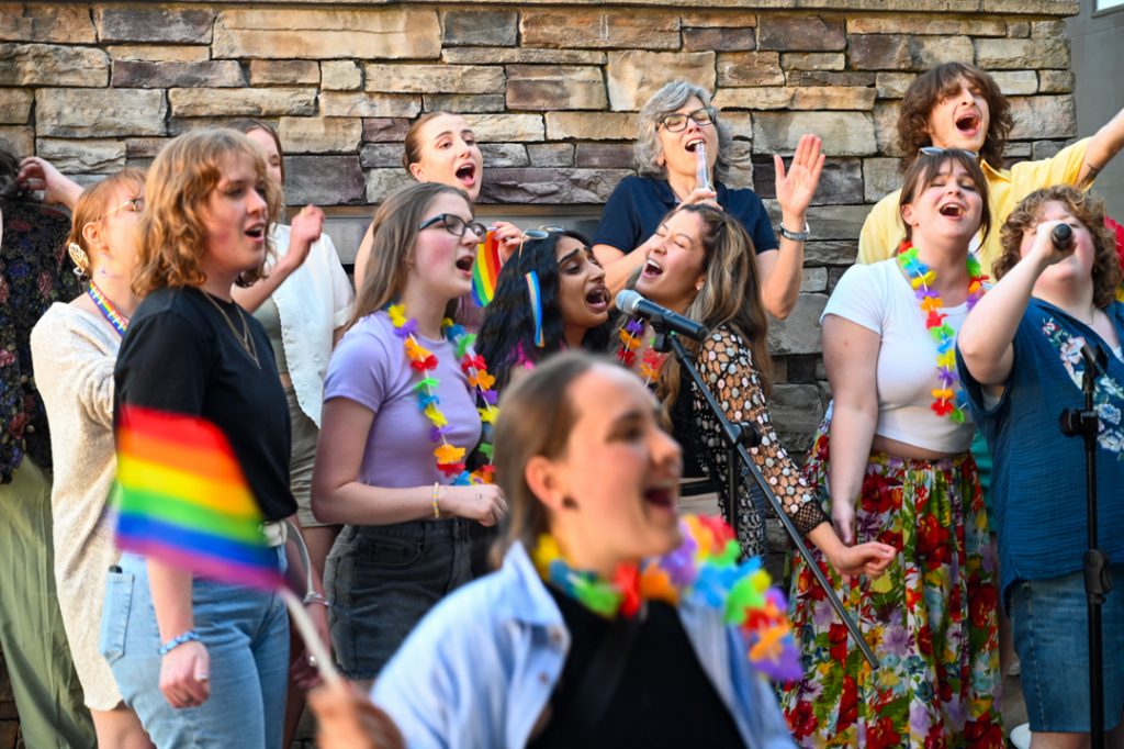 Photo of a group of people crowded onto an ourdoor stage, singing karaoke. Some hold rainbow flags or are wearing rainbow garlands