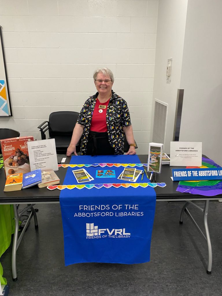 Photo of a person from another community organization standing behind another table with Friends of the Abbotsford Libraries branding
