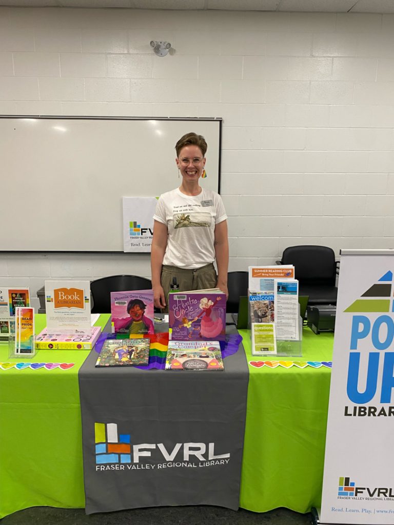 Photo of a person from another community organization standing behind another table with Fraser Valley Regional Library branding and a selection of books