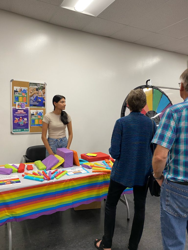 Photo of a person from another community organization standing behind another table, also with a rainbow flag tablecloth