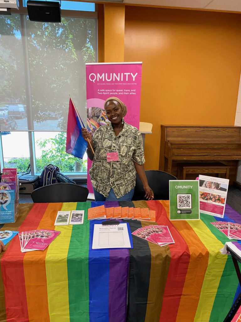 Photo of a person standing behind a table with a rainbow flag tablecloth, holding a flag, and in front of a sign that reads "Qmunity." There are brochures and signs on the table.