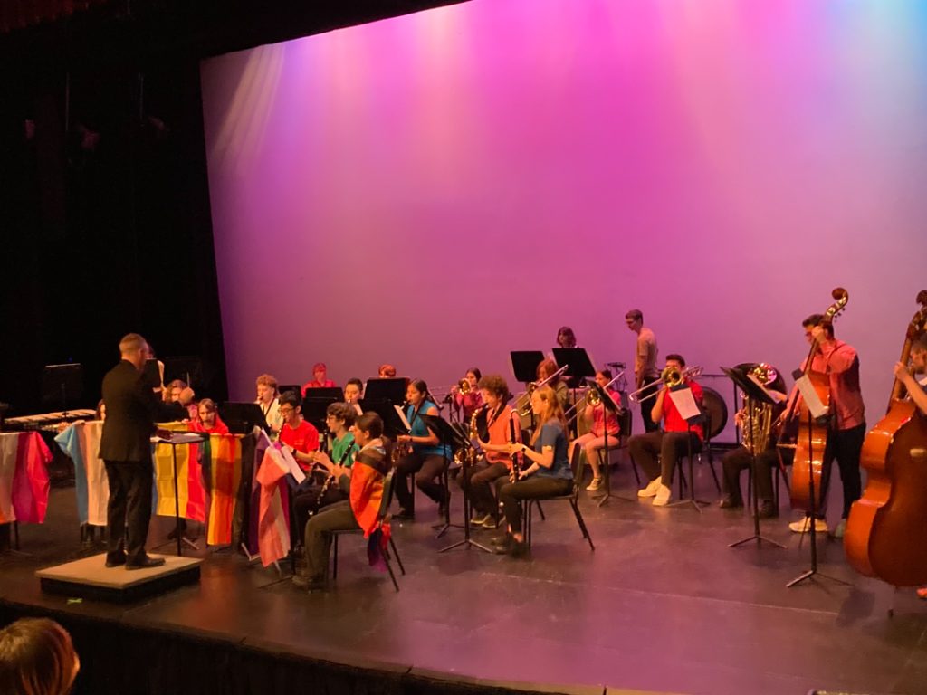 Photo of GW Graham's band performing on stage, with Pride flags hanging down from the front of several of the music stands.