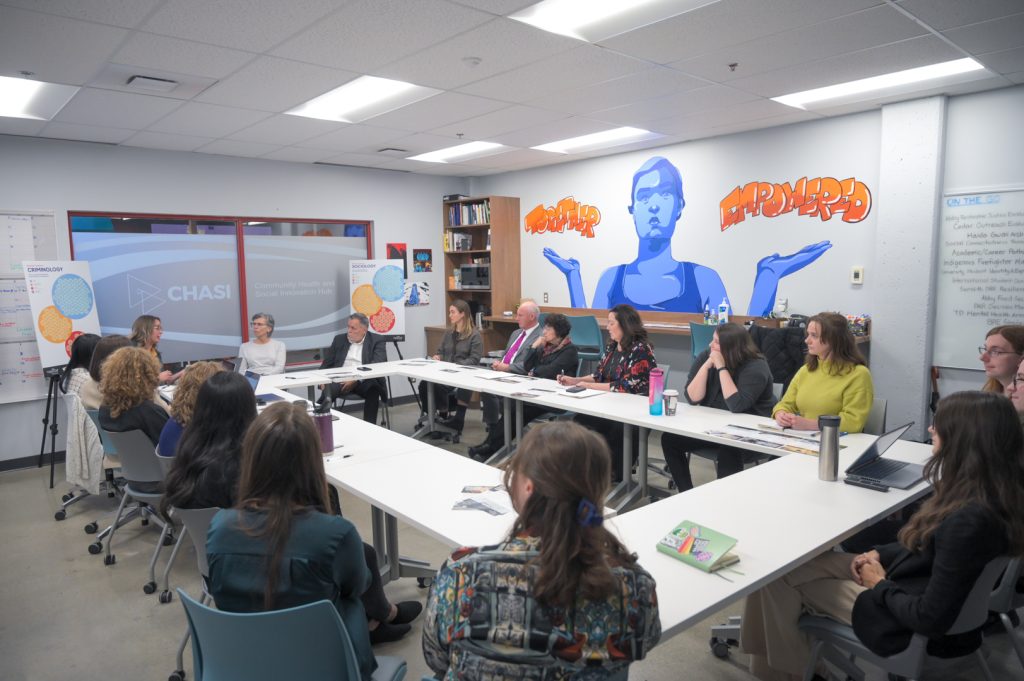 Wide shot photo of people surrounding a square of tables at CHASI. A wall shows a large mural of a blue-toned person with their hands held up like scales, holding the words "Together" and "Empowered." Everyone is looking at one person presenting to the group.