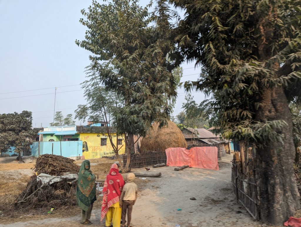Photo of a village with small buildings and large trees. Two women and a child walk away from the camera.