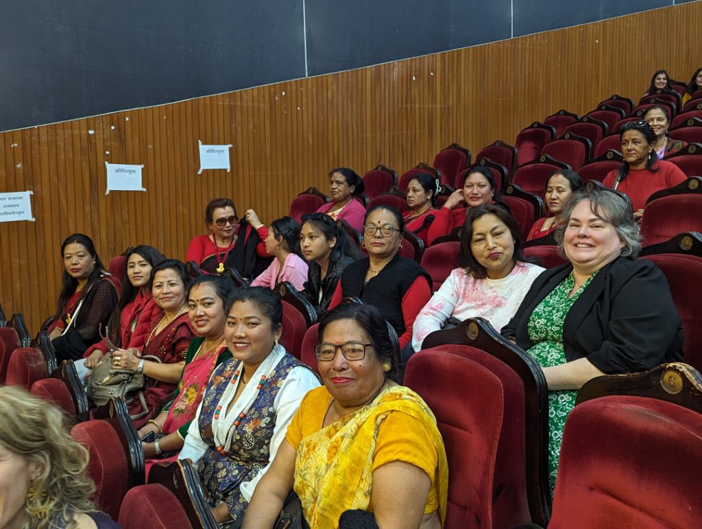 Photo of a large group of women, including Allie, sitting in theatre or lecture style seats an smiling for the camera.