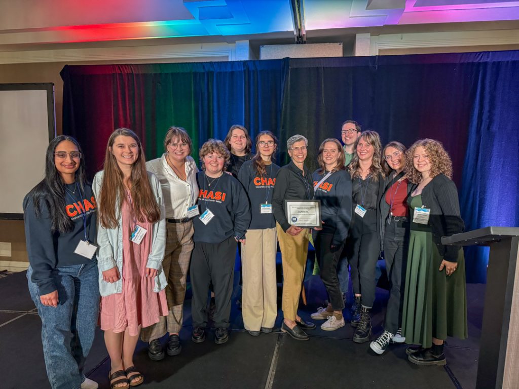 Photo of the 12 members of the CHASI team on stage posing for a photo. Dr. Martha Dow holds an award certificate. Four people are wearing CHASI-branded sweatshirts, and all have conference name badges on.