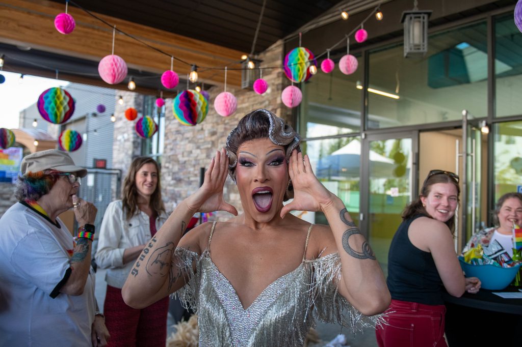 Photo of drag queen Kendall Gender posing for the camera, while Tea Dance attendees look on and smile. Colourful rainbow decorations hang from the ceiling.