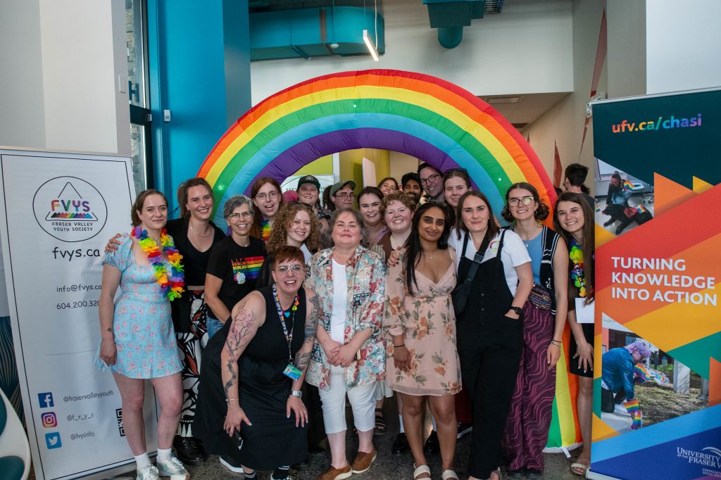 A large group of people are huddled together smiling at the camera. Behind them there is a large rainbow arch.