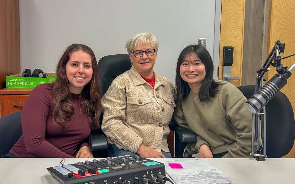 Photo of Ava Hedblom, Linda Pardy, and Mayu Ochi posing near a podcast microphone and mixer, smiling for the camera.