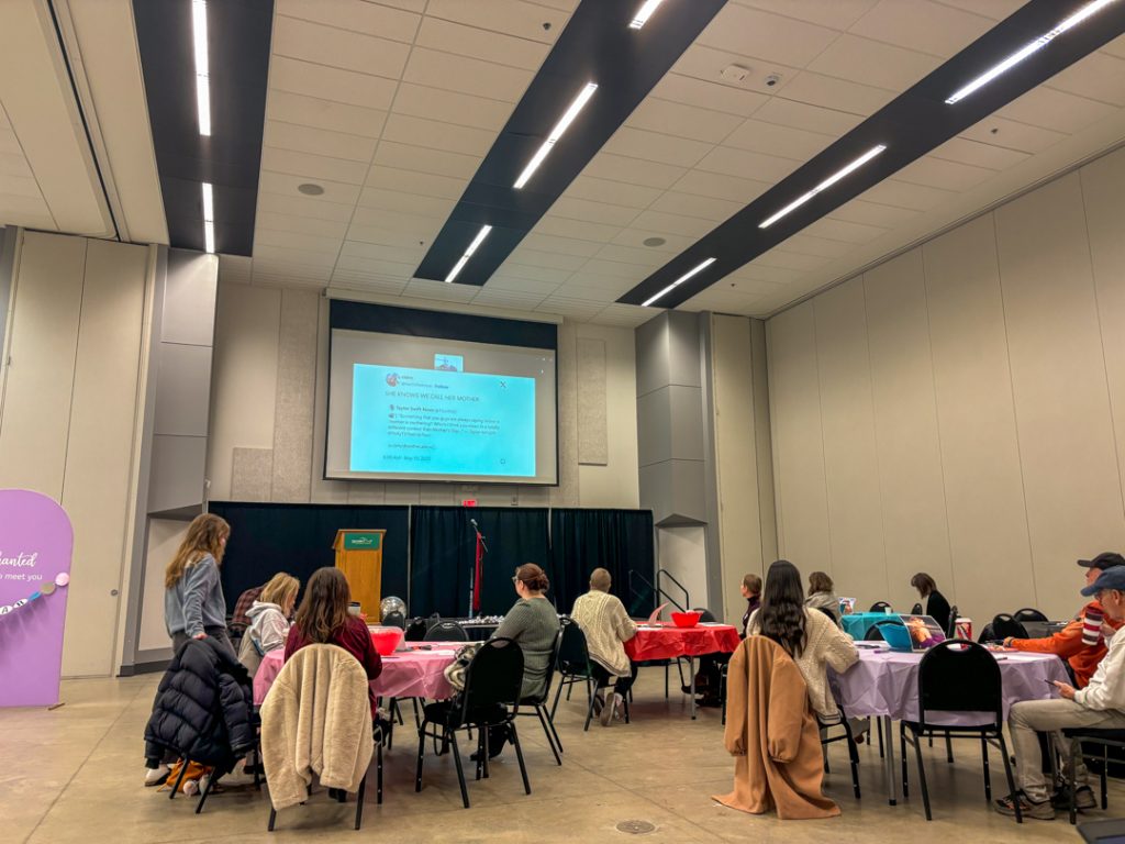 Conference attendees sit around five visible tables in front of a stage. They are looking up at a screen displaying a remote presenter.