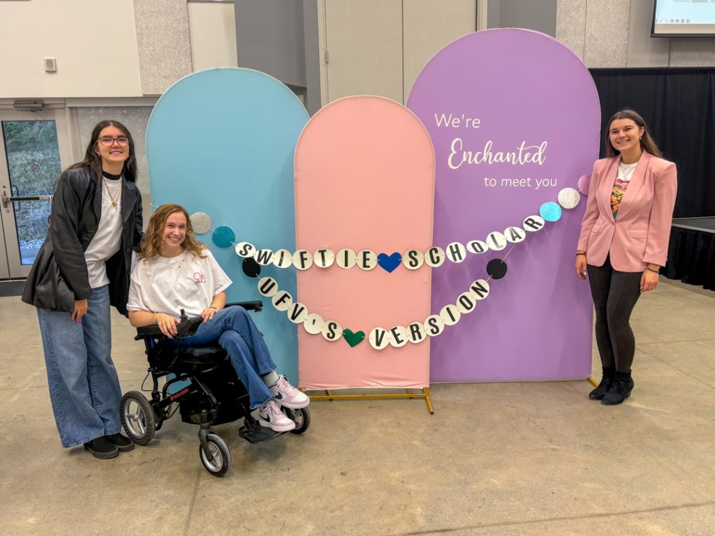 hree organizers of the Swiftie Scholar conference pose for a photo around a decoration in pastel colours with a large friendship bracelet banner. The friendship bracelet reads “Swiftie Scholar – UFV’s version.”