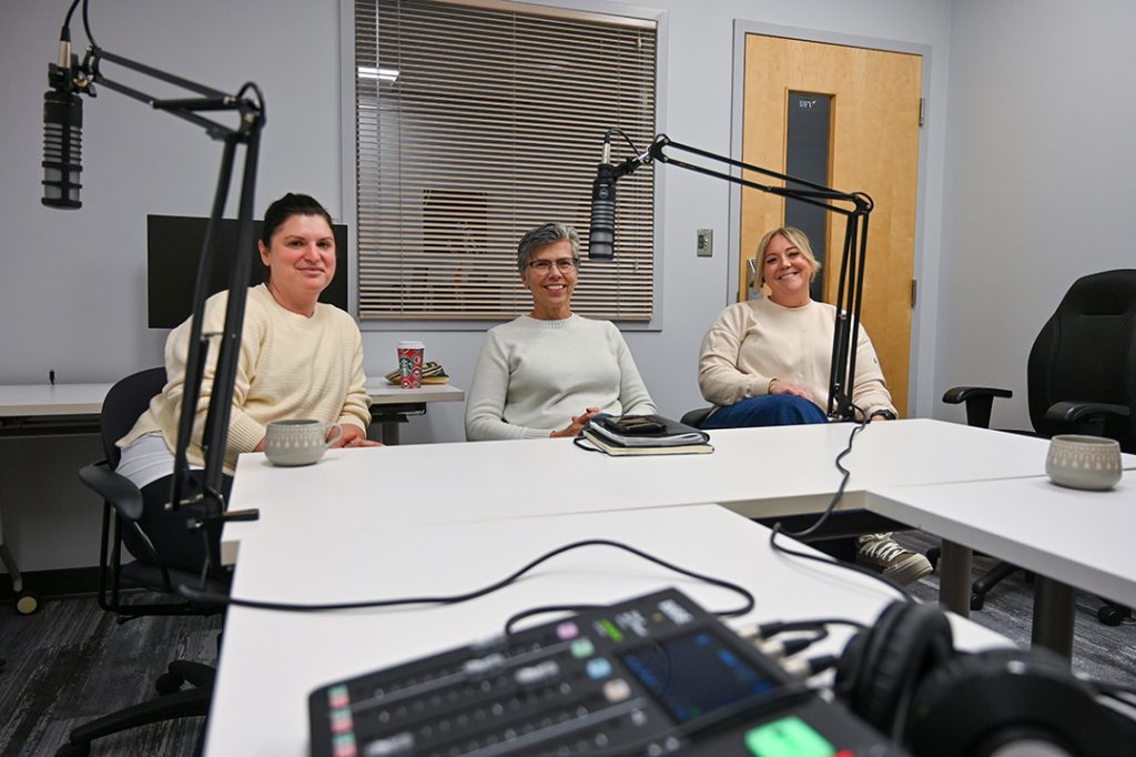 Photo of Iris Lesser, Martha Dow, and AmberJohnson posing near podcast microphones, smiling for the camera.