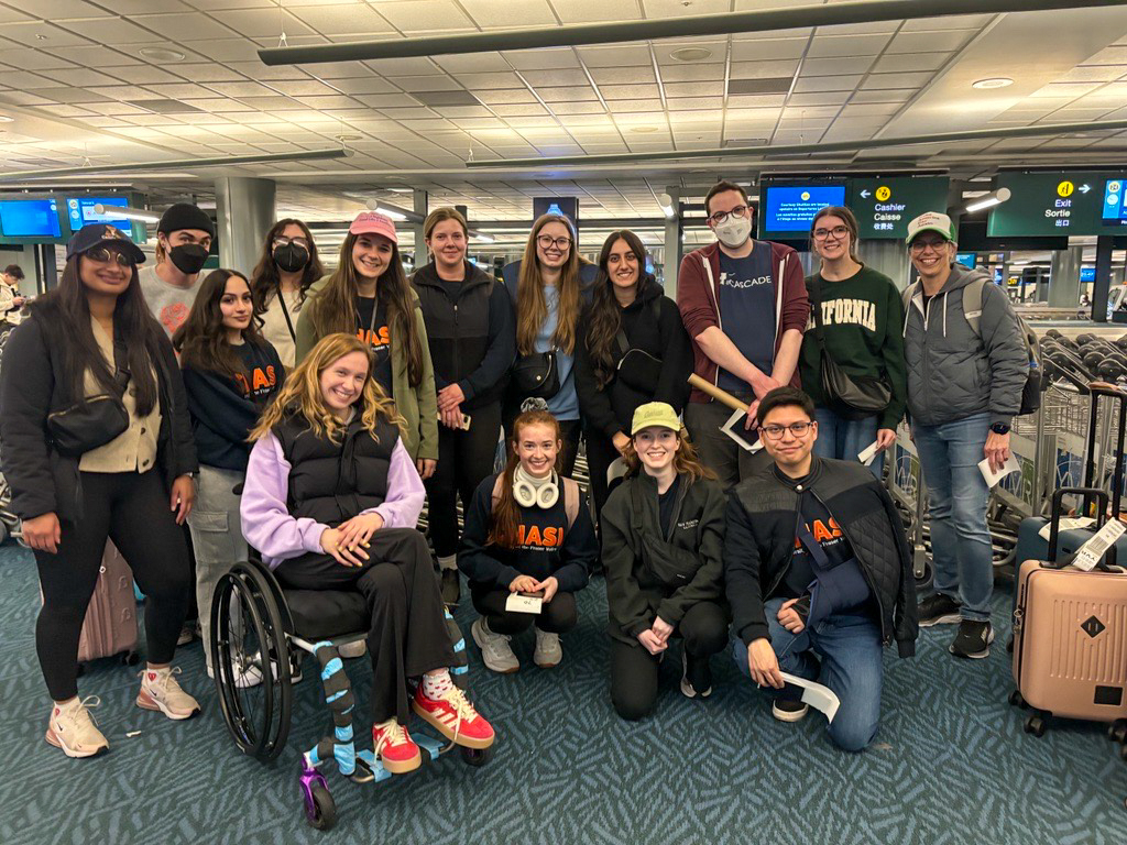 Group photo of 15 members of the CHASI team posing in an airport at the end of the trip home. Many are wearing navy blue sweatshirts with CHASI written on them in orange letters.