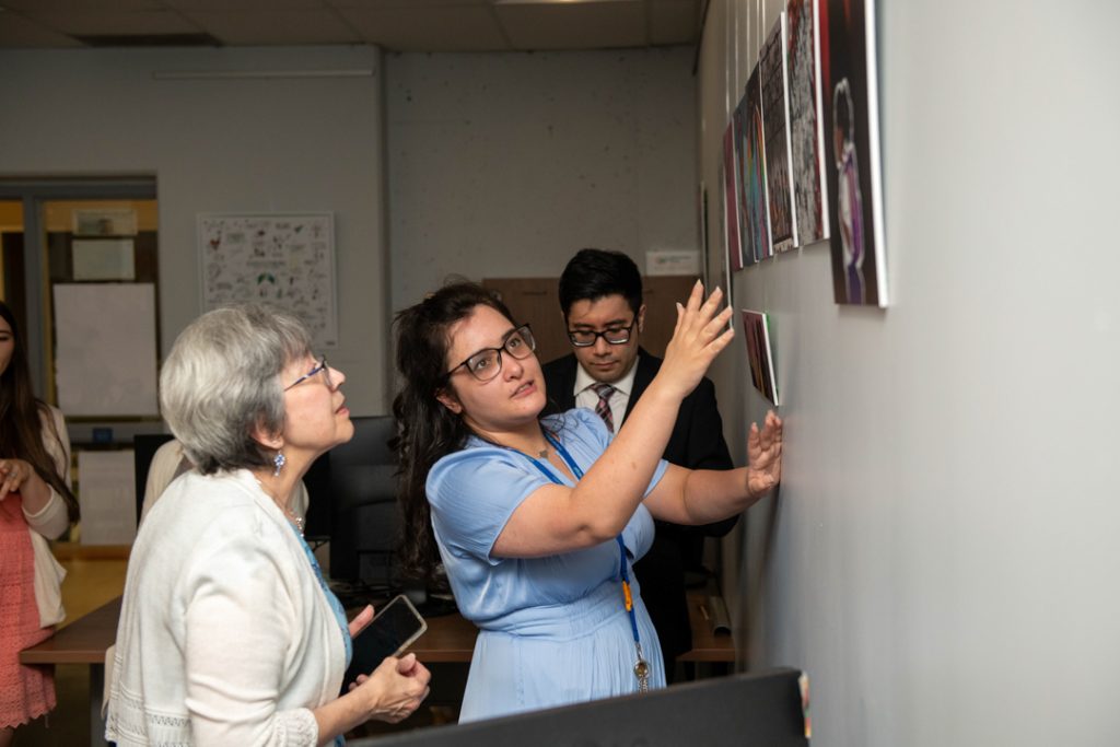Photo of Sharon Strauss showing her art, which is displayed on the wall, to a guest at the event.