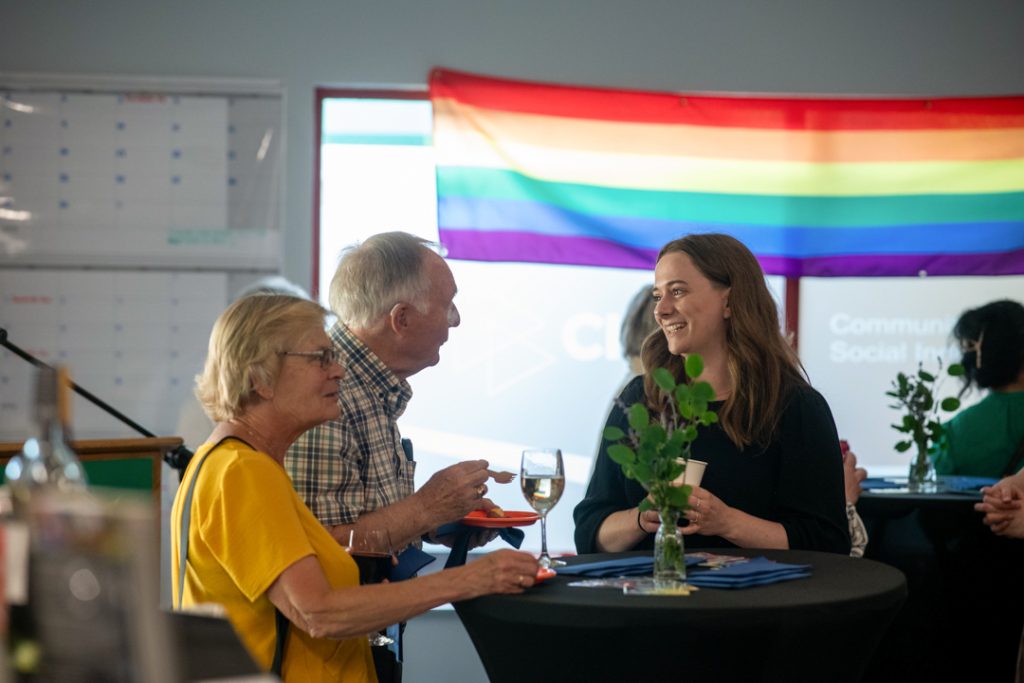 Photo of three people speaking and smiling around a tall table. The table has a plant centrepiece, and a large, bright pride flag hangs above a window behind them.