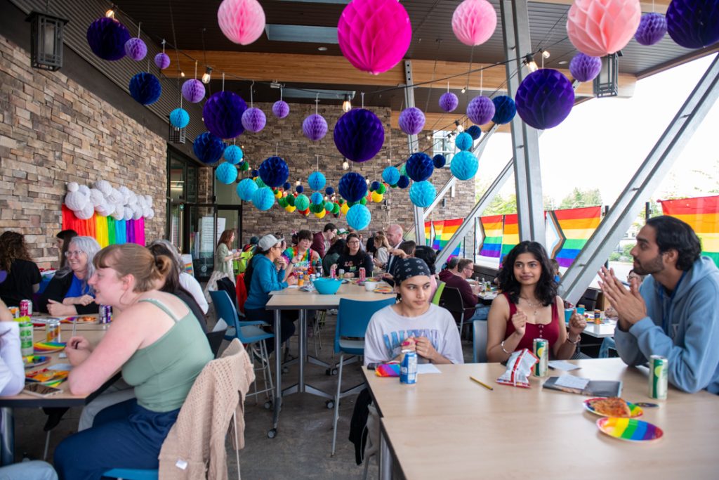Wide shot of the Tea Dance on the patio at UFV Abbotsford's K building. It is decorated with rainbow flags, hanging paper balloons, and rainbow plates and napkins. Each table has people around it, mingling and talking.