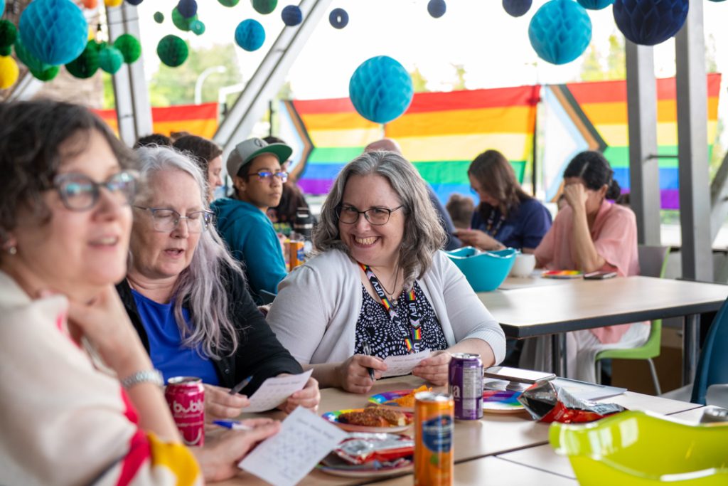 Attendees at the Tea Dance stand around a table, holding bingo cards. One smiles at the camera, while others look off camera towards the stage.