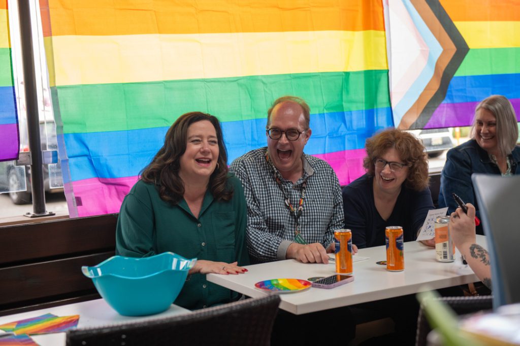 Group photo of attendees at the Tea Dance. They're sitting at a table with a large pride flag hanging behind them.