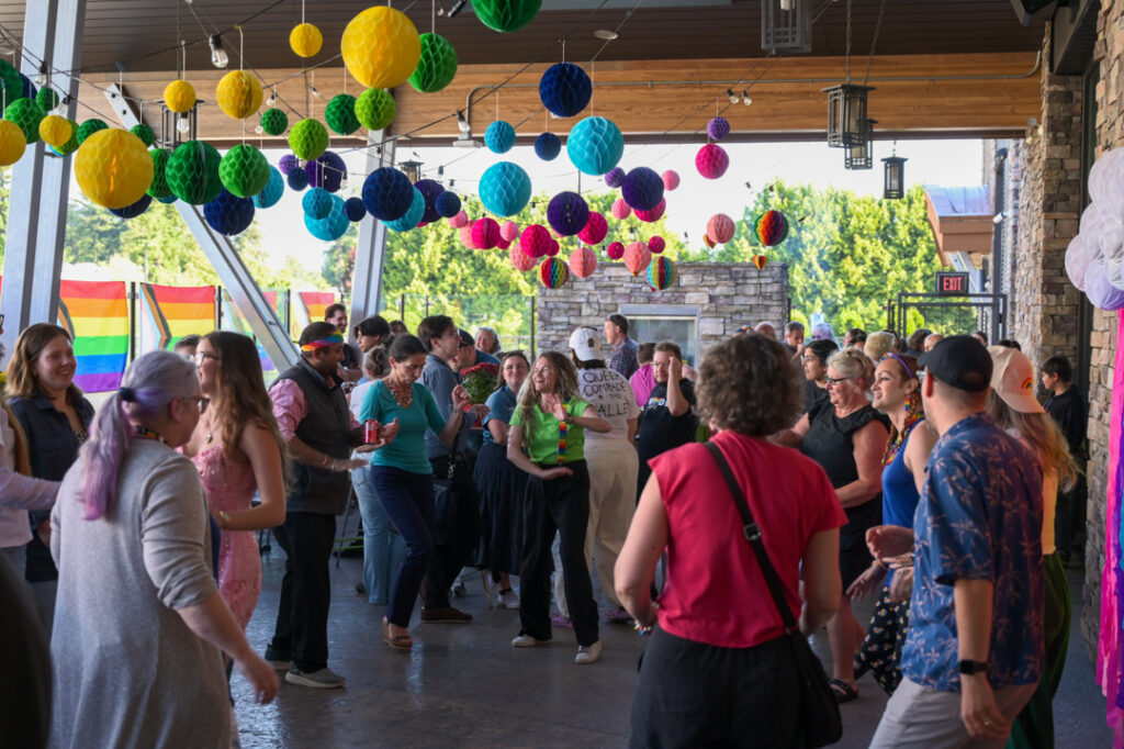 Photo of people dancing at the Tea Dance, forming a circle with a crowd watching and mingling outside. Colourful paper balloons hang above in rainbow colours, and Pride flags line the glass walls of the patio.