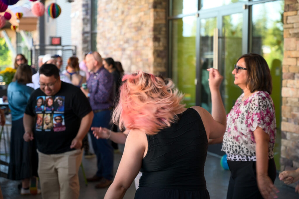 Photo of people dancing. In centre frame, a person with long pink hair is seen from behind, their hair flying in the wind as they move.