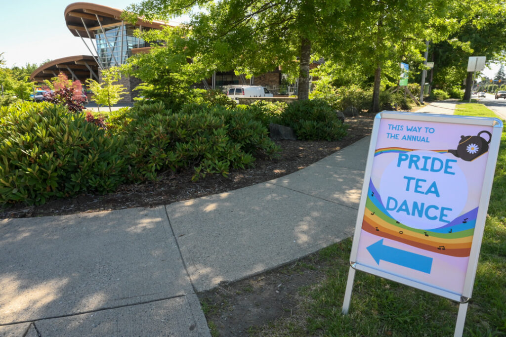Photo of a sign that reads "This way to the annual Pride Tea Dance." It has an arrow pointing towards UFV Abbotsford's K building, which is visible in the background. It is a lovely day!