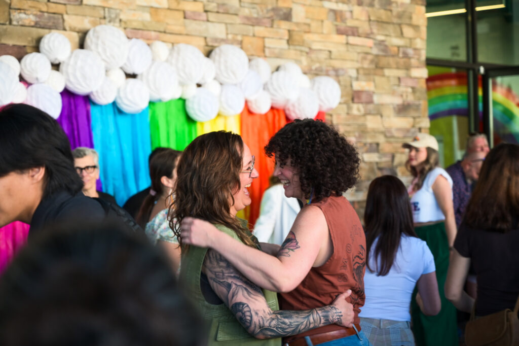 Photo of two people holding each other in a slow dance with huge smiles on their faces as they look into each others' eyes. In the background is a a wall of rainbow decorations.