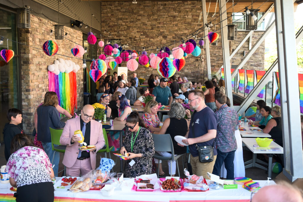 Crowd shot of the Tea Dance. People are happy, talking, serving themselves food from a table, and dancing. Bright, rainbow colour decorations and Pride flags hang from the walls and ceiling.