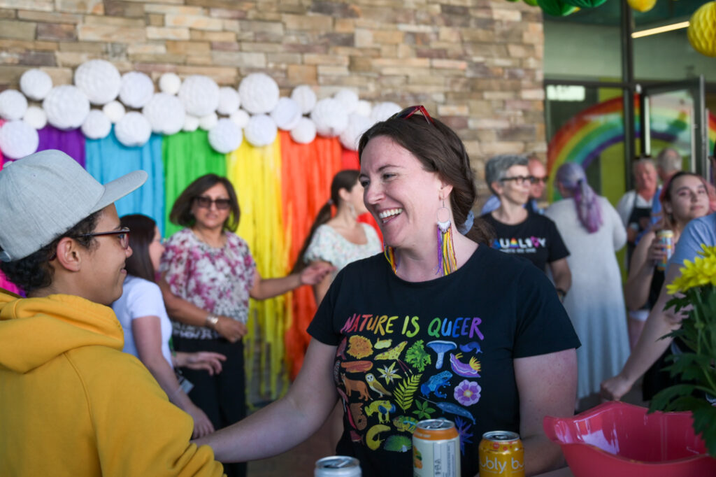 Photo of a person at the Tea Dance, with colourful rainbow decorations behind her. She is smiling, as she speaks, and wearing a t-shirt that says "nature is queer" with rainbow-coloured pictures of plantlife.
